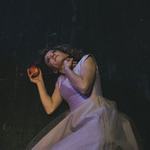 Młoda kobieta w białej sukience leży na deskach sceny. W dłoni trzyma jabłko.