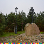 Głaz z metalowym krzyżem prawosławnym. Na drugim planie trzy drewniane krzyże, metalowa latarnia i rosnące drzewa.