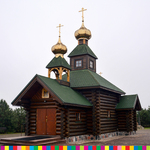 Drewniana cerkiew z dwiema wieżami uwieńczonymi złotymi kopułami i krzyżami prawosławnymi. Na drugim planie rosną drzewa i krzewy.