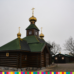 Drewniana cerkiew z trzema wieżami ze złotymi kopułami i krzyżami. Na drugim planie zabudowania i rosnące drzewa.