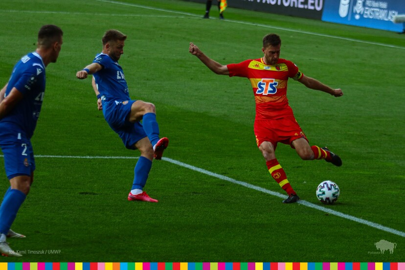 Zawodnik w żółto czerwonym stroju kopie piłkę obok zawodnika w niebieskim stroju