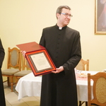 Na zdjęciu widoczny kapłan trzymający dokument oprawiony w grube ramy. Obok niego stoi po lewej abp Tadeusz Wojda. W tle widoczne stół i krzesła. Na ścianie widać obraz Jezusa Miłosiernego
