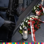 Wieńce z biało-czerwoną wstęgą leżące pod tablicą upamiętniającą zamordowanych Żołnierzy Wyklętych. Z lewej strony widoczne dłonie w rękawicach trzymające broń