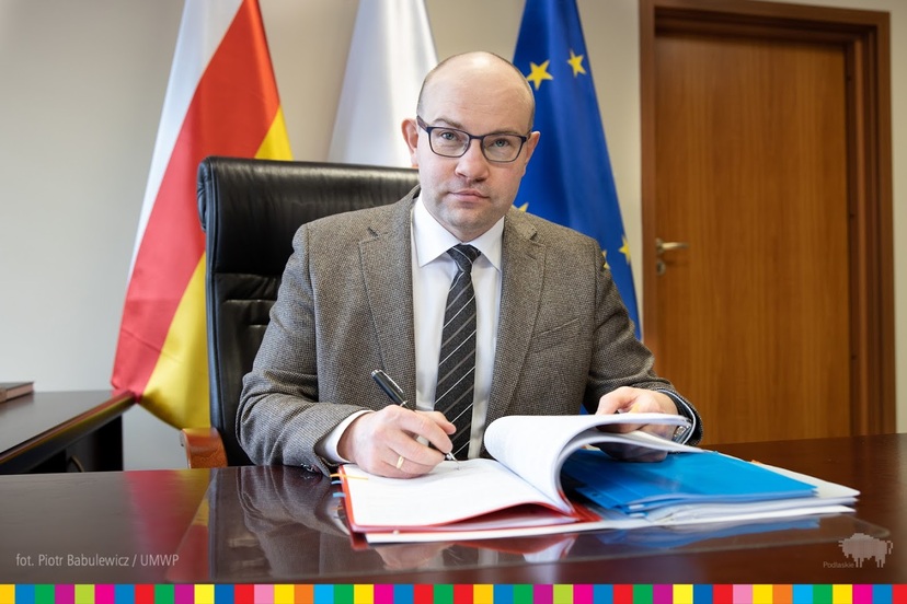 Marszałek Artur Kosicki podpisuje umowę. W tle flagi Województwa Podlaskiego, Polski oraz UE.