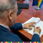 Wicemarszałek Marek Olbryś podpisuje umowę na wykonanie mikroinstalacji Wąsosz. W tle widoczna niebieska flaga UE oraz biało-czerwono-żółto-niebieska