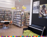 Wnętrze biblioteki. Półki z ksiażkami, stolik i krzesła. Instrument muzyczny.