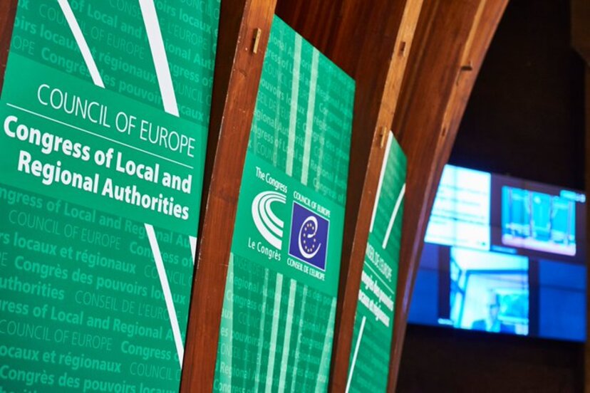 Po lewej zielone plansze z napisem w języku angielskim: Kongres Władz Lokalnych i Regionalnych Europy. Po prawej ekran monitora z uczestnikami.