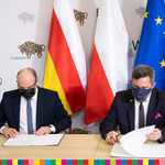 Artur Kosicki i Radosław Dobrowolski podpisują umowę siedząc za stołem. Za nimi flagi: województwa, Polski, Unii Europejskiej