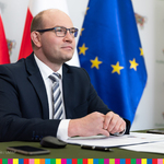 Profil marszałka Kosickiego siedzącego za stołem. W tle flagga unijna i polska