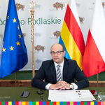 Marszałek Kosicki siedzi za stołem prezydialnym. Za nim ścianka promocyjna województwa, laska marszałkowska oraz flaga unijna, województwa podlaksiego oraz Polski