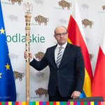 Marszałek Kosicki z laską marszałkowską w prawej dłoni, na tle flag: europejskiej, polskiej i wojewódzkiej