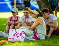 Grupa ludzi siedzi na trawie. Prze nimi stoi tabliczka z rysunkiem jak postać szepcze coś do ucha.