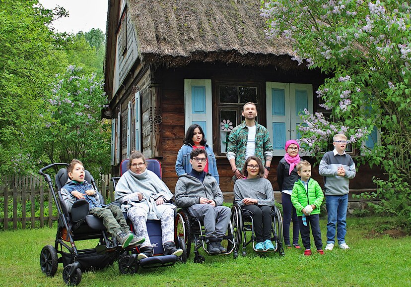 Grupa dzieci z niepełnosprawnością wraz z Przemysławem Kossakowskim przed drewnianą chatą