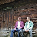 Przemysław Kossakowski wraz z innym mężczyzna siedzą na ławce przed drewniana chatą