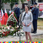 Kobieta przemawia przez mikrofon. Obok leżą wieńce oraz stoją flagi biało-czerwone. W tle stoją wartownicy