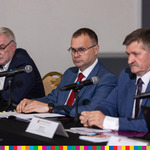 Trzech mężczyzn w garniturach siedzi przy stole z mikrofonami