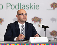 Artur Kosicki siedzi za stołem konferencyjnym. Przed nim mikrofon. Za nim ścianka z logotypami Podlaskiego oraz napis Podlaskie.