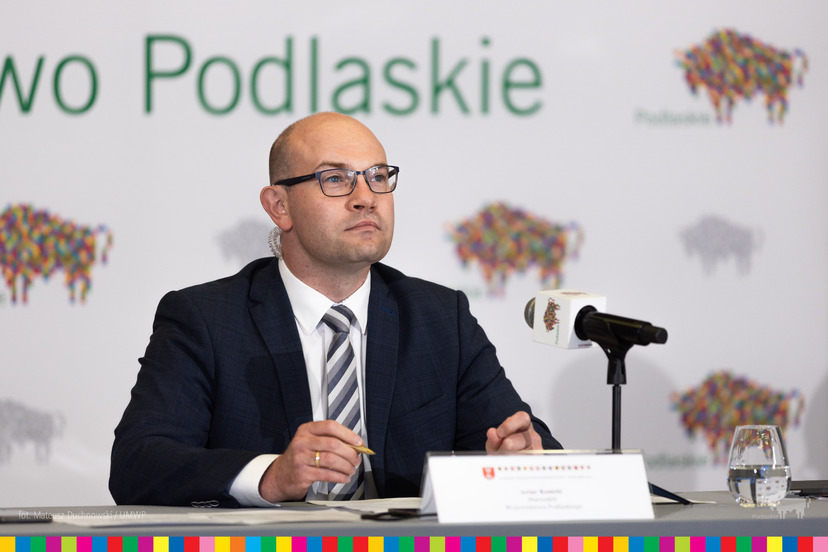 Artur Kosicki siedzi za stołem konferencyjnym. Przed nim mikrofon. Za nim ścianka z logotypami Podlaskiego oraz napis Podlaskie.
