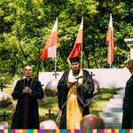 Trzy osoby duchowne stoją na zdjęciu. Za nimi mogiły powstańców oraz flagi biało-czerwone
