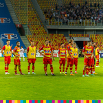 Grupa piłkarzy w żółto-czerwonych strojach stoi na boisku. W tle widoczne trybuny