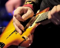 Zbliżenie na gitarę elektryczną, na której gra mężczyzna, dłonie z kostką nad strunami gitary