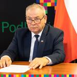 Wicemarszałek marek Olbryś siedzi przy stole podczas konferencji. Za nim znajduje się flaga Polski oraz Unii Europejskiej