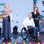 Wiesłąwa Burnos, mężczyzna na wózku inwalidzkim oraz stojąca obok niego kobieta na scenie podczas wręczenie nagród