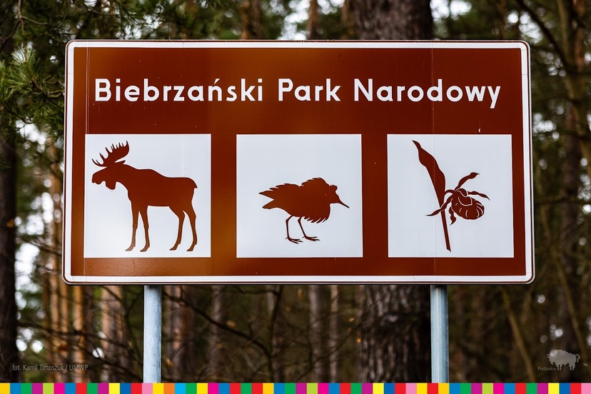 Tablica informacyjna z napisem Biebrzański Park Narodowy. Poniżej znaki: łoś, ptak i owad.