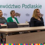 Trzy kobiety siedzą na sali, za nimi widoczny baner z logo Województwa Podlaskiego 