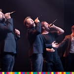 Mężczyźni śpiewają na scenie trzymając mikrofony w górze