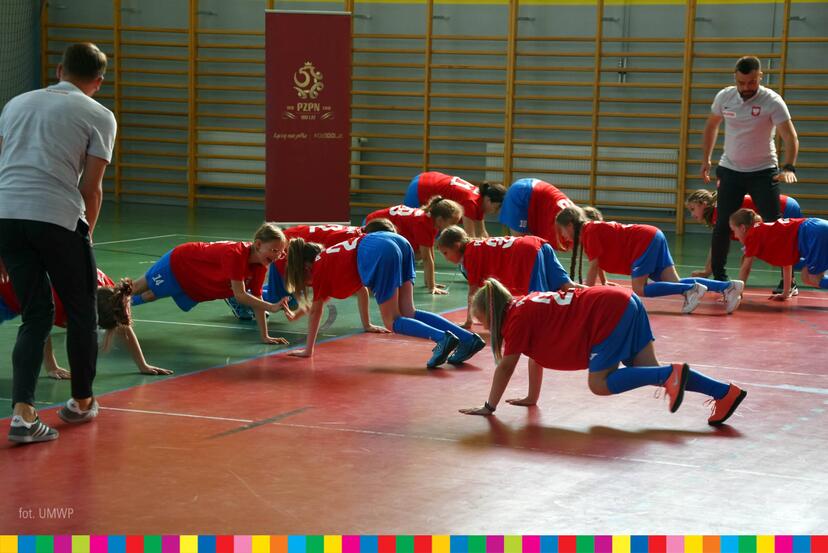 Dzieci w czerwonych koszulkach i niebieskich spodenkach ćwiczą na sali gimnastycznej