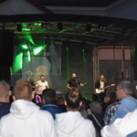 Na scenie grają mężczyźni na gitarach. Przed sceną stoją tłumy
