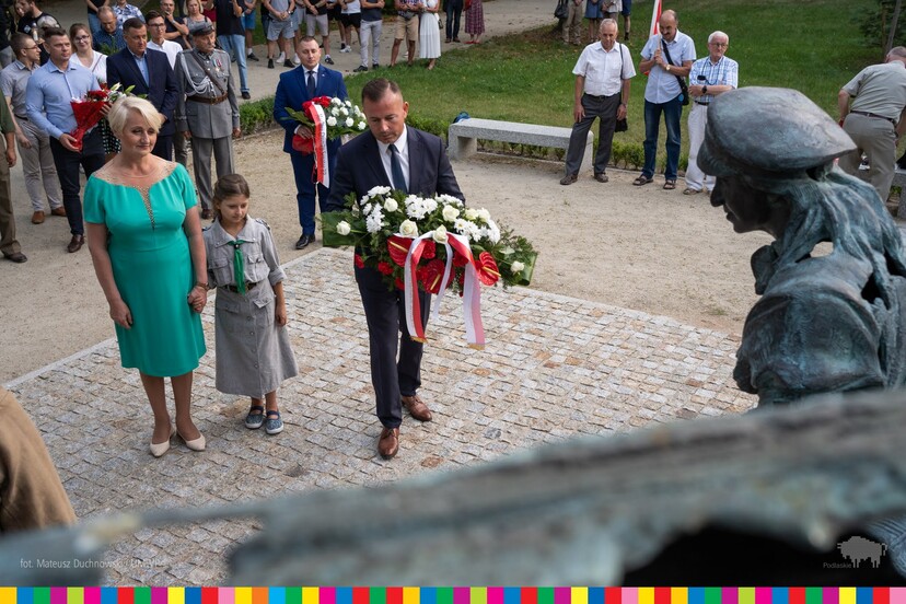 Mężczyzna trzyma kwiaty przewiązane biało-czerwoną wstążką. Po lewej stronie stoi jasnowłosa kobieta, za rękę trzyma dziewczynkę w mundurze harcerskim. Za nimi stoją pozostali uczestnicy wydarzenia.