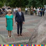 Przed Pomnikiem Katyńskim stoją kobieta w niebieskiej sukience, mężczyzna w garniturze, w tle stoją uczestnicy wydarzenia.