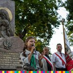 Przy Pomniku Katyńskim w Parku Konstytucji 3 Maja stoi dziewczynka w mundurze harcerskim, trzyma mikrofon przy ustach. Po prawej w tle stoi trzech mężczyzn z przewiązanymi szarfami biało-czerwonymi, jeden z nich trzyma sztandar.