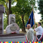 Przed Pomnikiem Katyńskim w Parku Konstytucji 3 Maja stoi trzech mężczyzn przewiązanych szarfami biało-czerwonymi. Mężczyzna stojęcy pośrodku trzyma w rękach granatowy sztandar.