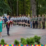 Kobieta w niebieskiej sukience i mężczyzna w garniturze idą w kierunku pomnika. Mężczyzna w rękach trzyma wiązankę kwiaty w kolorach biało-czerwonych. Po prawej w rzędzie stoją mężczyźni w mundurach, harcerze i pozostaki uczestnicy wydarzenia.