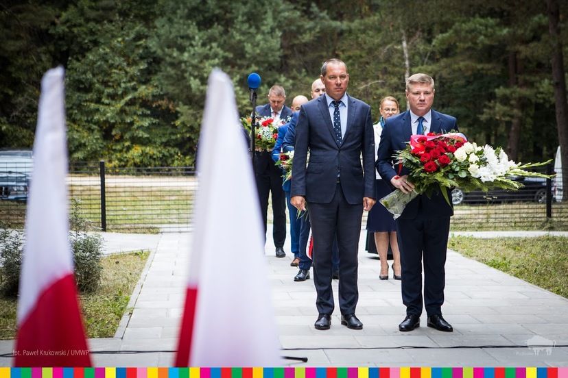 Dwóch mężczyzn w garniturach, jeden z nich trzyma w rękach kwiaty w kolorach biało-czerwonych. Za nimi inni uczestnicy wydarzenia trzymają kwiaty.