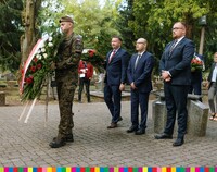 Żołnierz trzyma wieniec z kwiatów biało-czerwonych, za nim stoją Artur Kosicki, Sebastian Łukaszewicz, inni uczestnicy wydarzenia.