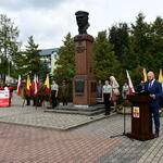 Pomnik J. Piłsudskiego z obu stron stoją harcerze, z lewej strony przy flagach mężczyźni w mundurach, po prawej mężczyzna z mikrofonem za nim inni uczestnicy wydarzenia. 